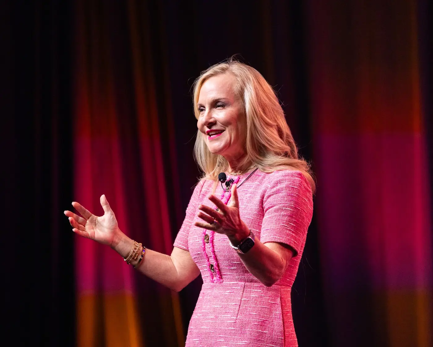 A speaker in a pink jacket addresses the audience onstage.