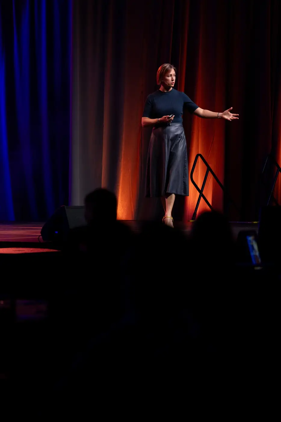 A speaker gestures on a dimly lit stage beside the forum screen.